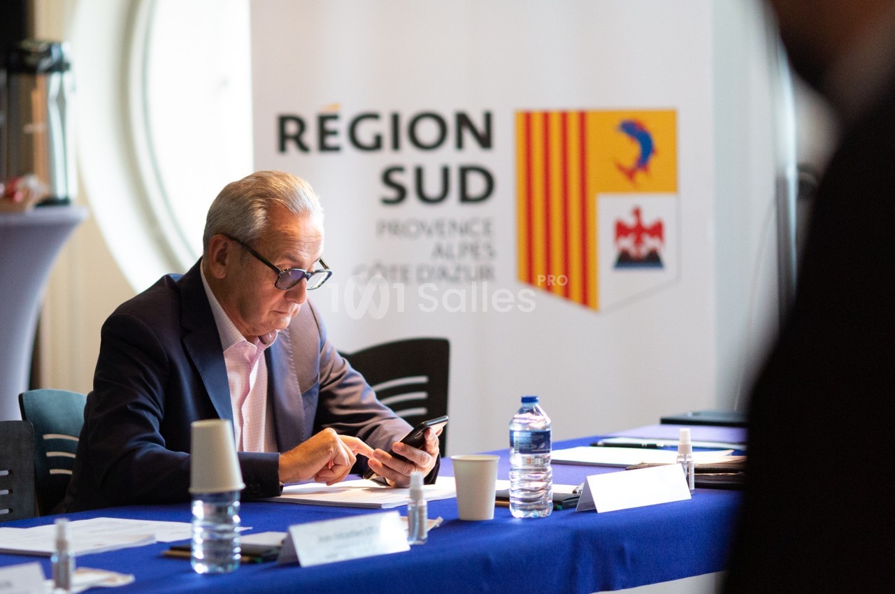 Un homme consulte son téléphone lors d'une réunion, avec un panneau ’Région Sud’ en arrière-plan.