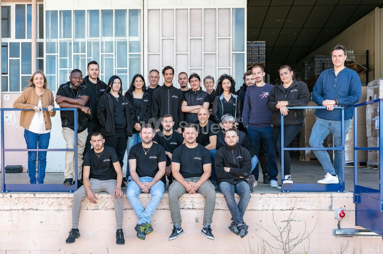Un groupe de personnes posant devant un bâtiment industriel avec des portes vitrées et des palettes en arrière-plan.