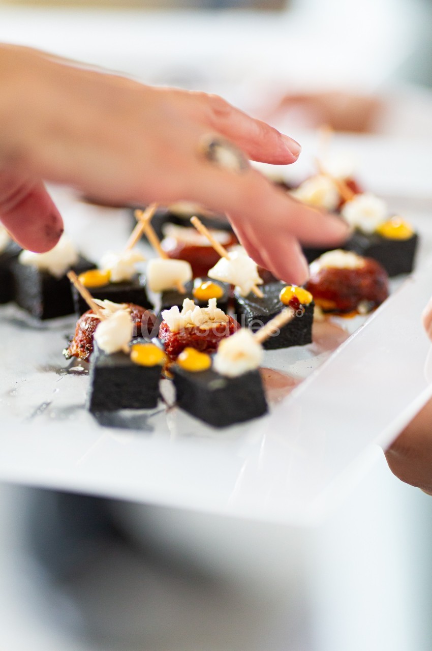 Des mains disposant des amuse-bouches sur un plateau, composés de morceaux noirs, garnis de fromage et de tomates.