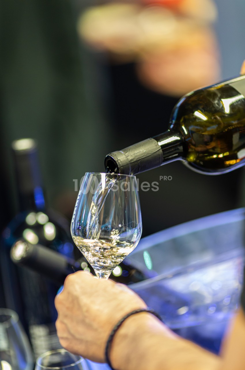 Une main verse du vin blanc dans un verre transparent, avec des bouteilles en arrière-plan flou.