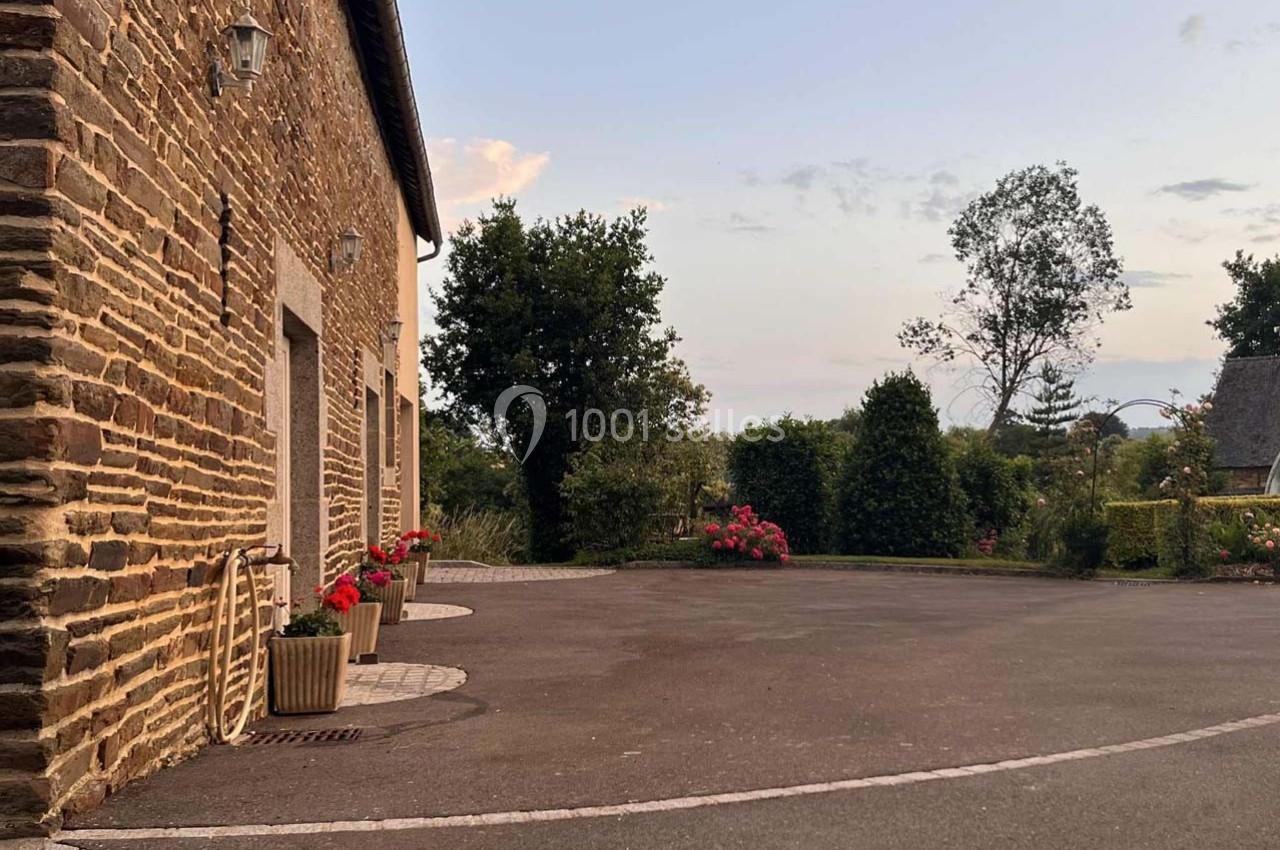 Cour pavée devant une maison en pierre avec des pots de fleurs, entourée de verdure et d'arbres au crépuscule.