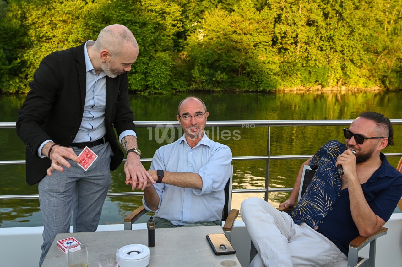 Un magicien réalise un tour de cartes pour deux hommes assis sur une terrasse au bord d'une rivière.