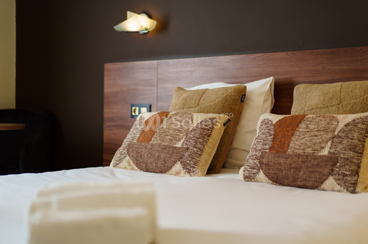 Lit double avec tête de lit en bois, coussins décoratifs beige et marron, et mur sombre en arrière-plan.