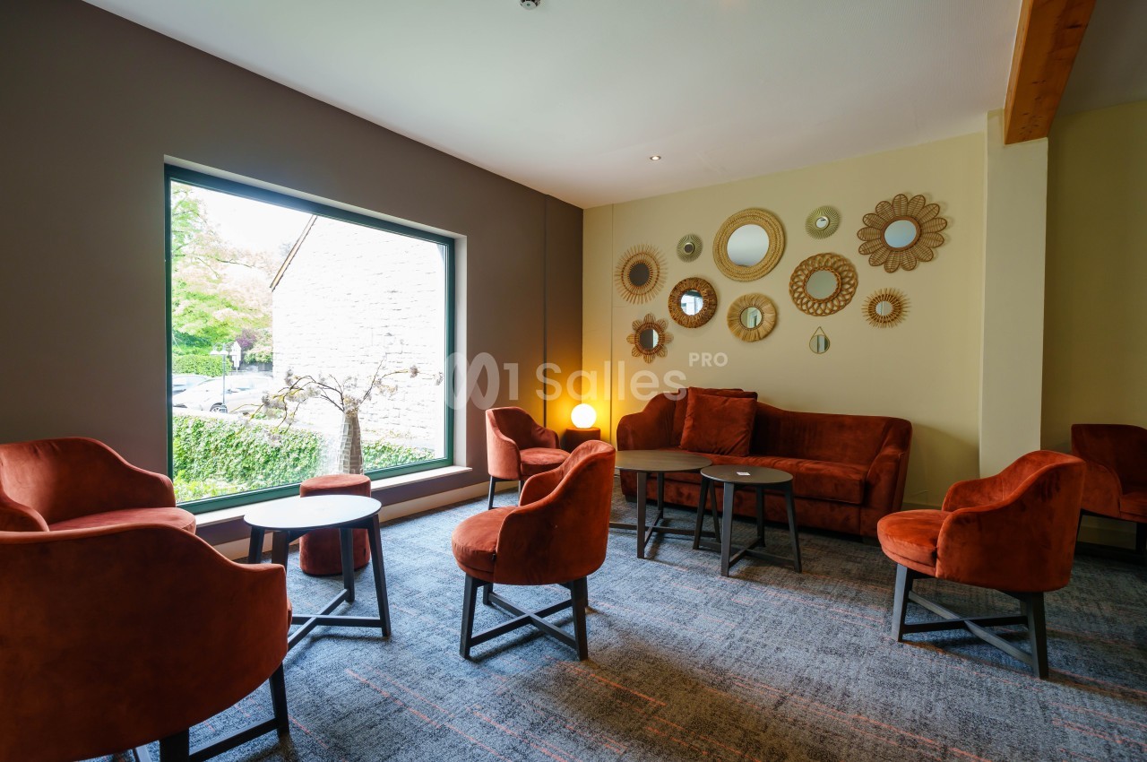 Salon lumineux avec fauteuils et canapé orange, tables basses noires, miroirs décoratifs et grande fenêtre donnant sur un…