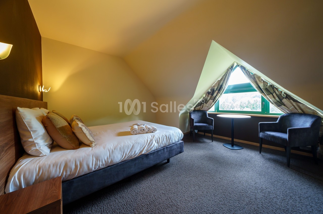Chambre mansardée avec lit simple, deux fauteuils, table ronde et fenêtre triangulaire offrant une vue sur la nature.