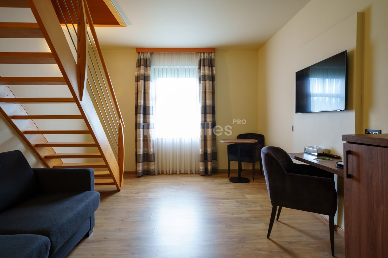Salon lumineux avec parquet, canapé, escalier en bois, bureau avec chaise, télévision murale et fenêtre avec rideaux.