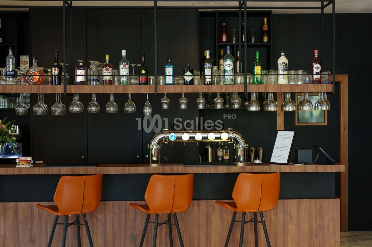 Bar moderne avec comptoir en bois, tabourets orange, étagères de bouteilles et verres suspendus.