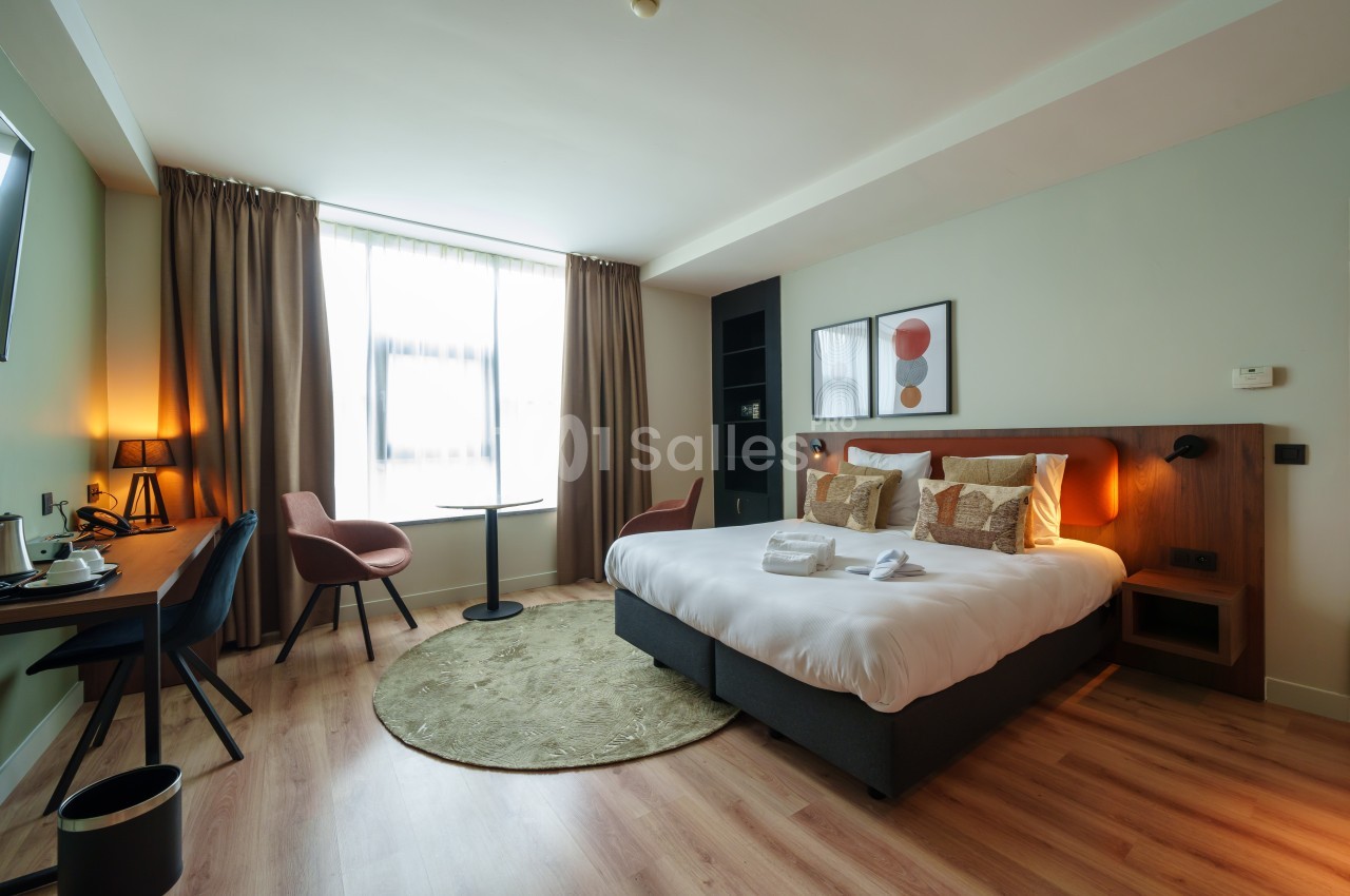 Chambre d'hôtel lumineuse avec lit double, bureau, fauteuils, table ronde et grande fenêtre avec rideaux.