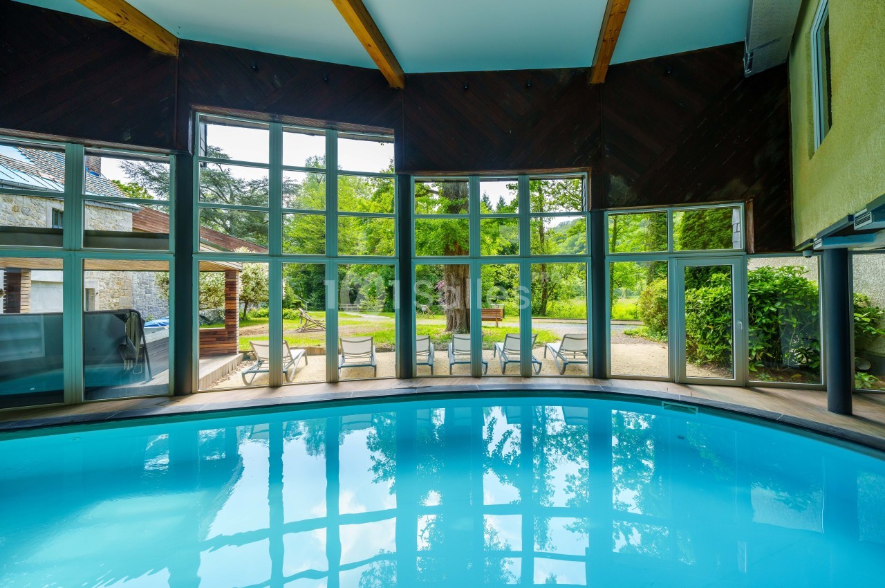 Piscine intérieure avec grandes baies vitrées offrant une vue sur un jardin verdoyant et des chaises disposées à l'extérieur.