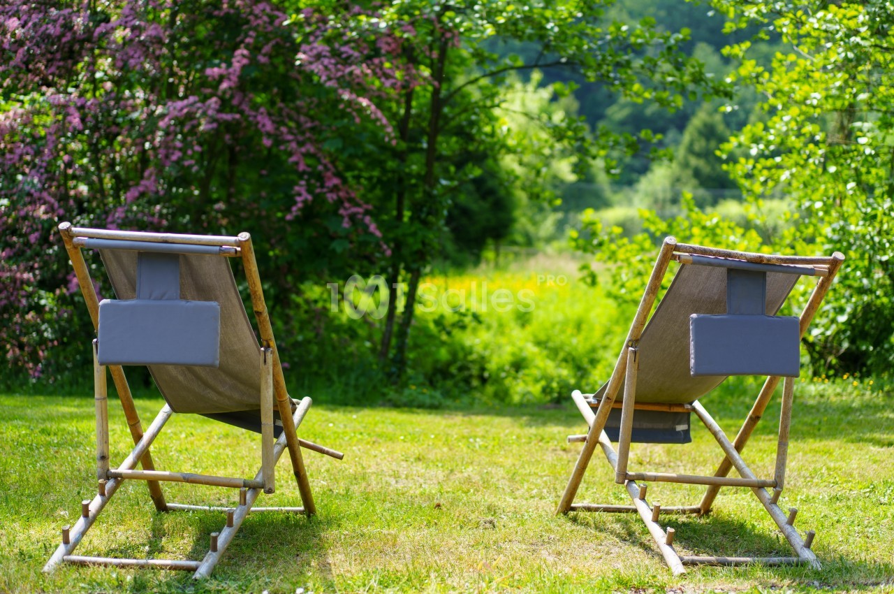 Deux chaises longues en bois et tissu gris installées sur une pelouse, entourées de végétation verdoyante et d'arbustes…