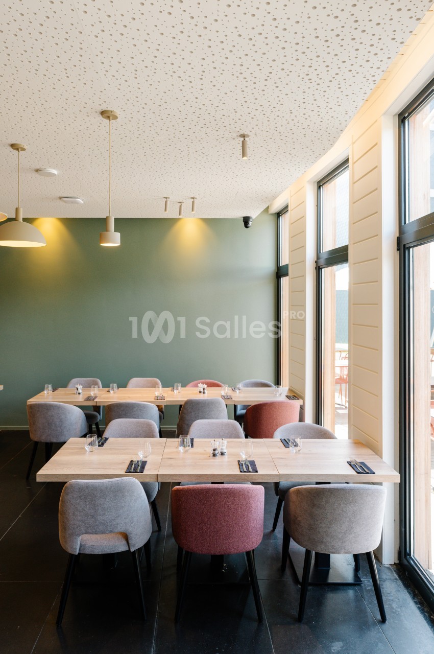 Salle de restaurant moderne avec tables en bois, chaises colorées et lumière naturelle entrant par de grandes fenêtres.