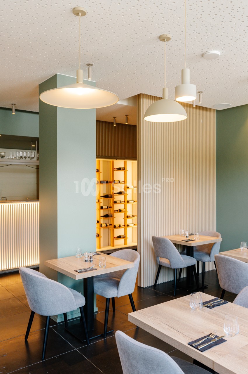 Salle de restaurant moderne avec tables en bois clair, chaises grises et éclairage suspendu, vue sur une cave à vin vitrée.