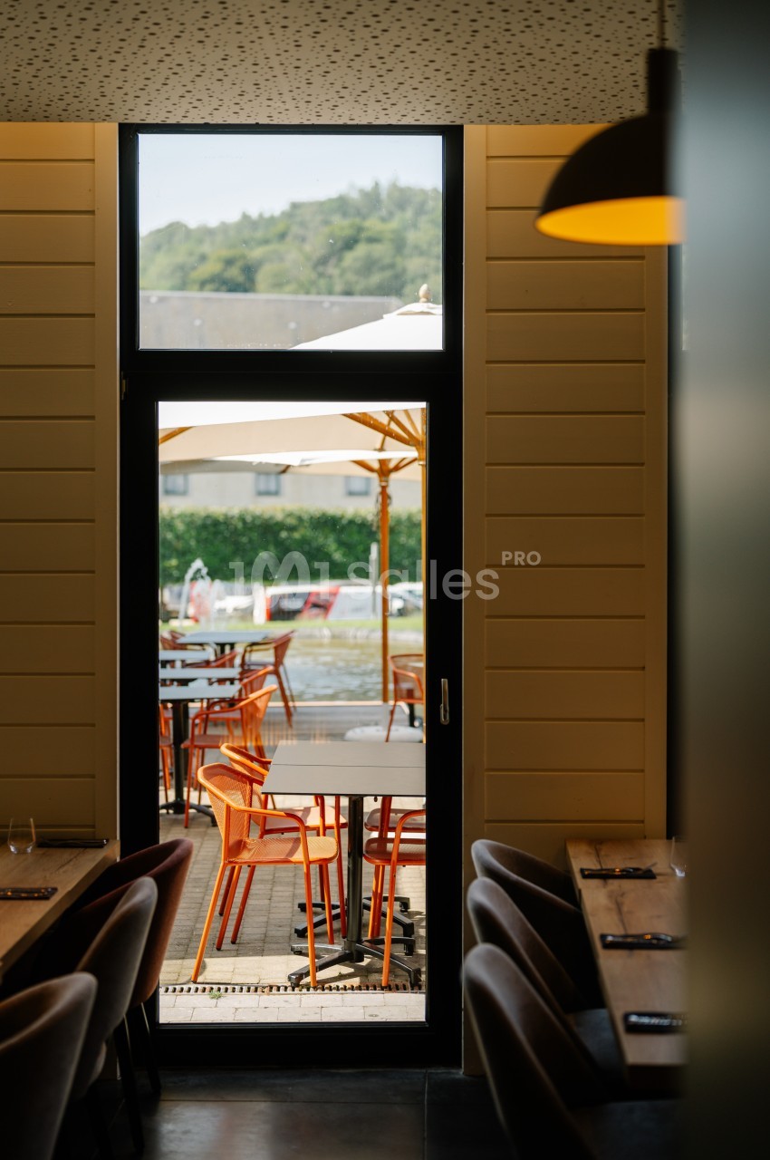 Vue depuis l'intérieur d'un restaurant sur une terrasse extérieure avec des tables, chaises orange et parasols.