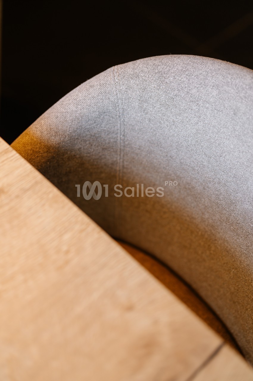 Chaise en tissu gris clair partiellement visible près d'une table en bois clair, éclairée par une lumière douce.