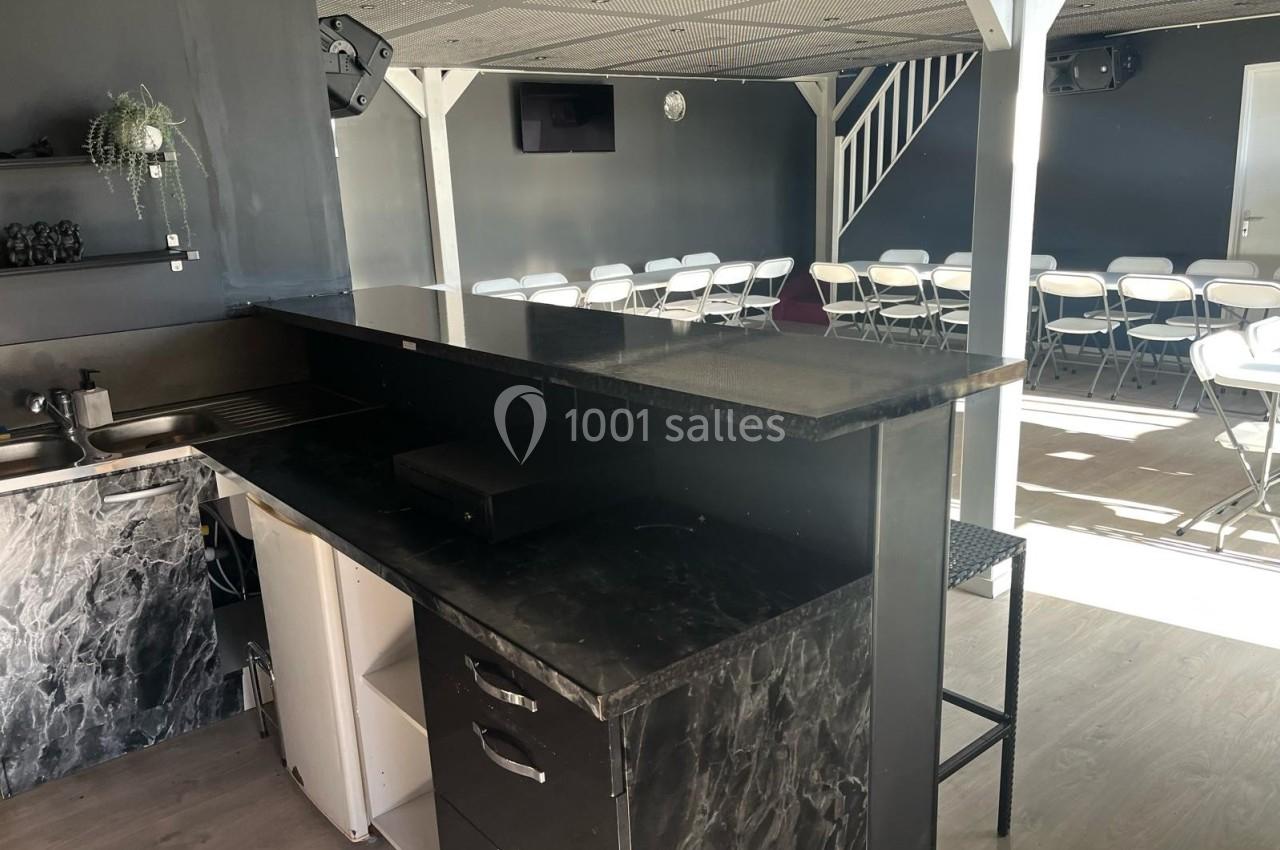 Salle avec un comptoir noir, un évier, des chaises blanches disposées en rangées et un escalier au fond.
