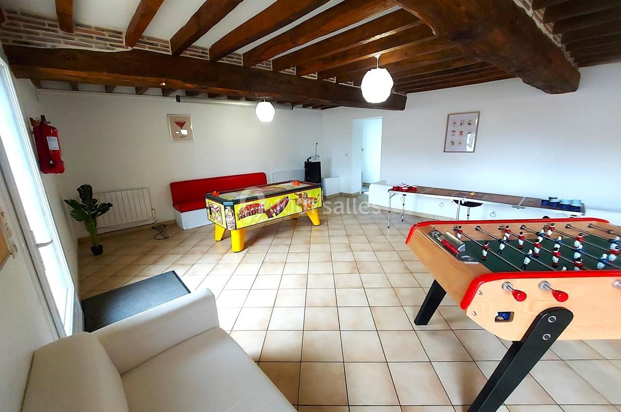 Salle de jeux avec baby-foot, flipper, canapé, murs blancs et poutres apparentes au plafond.