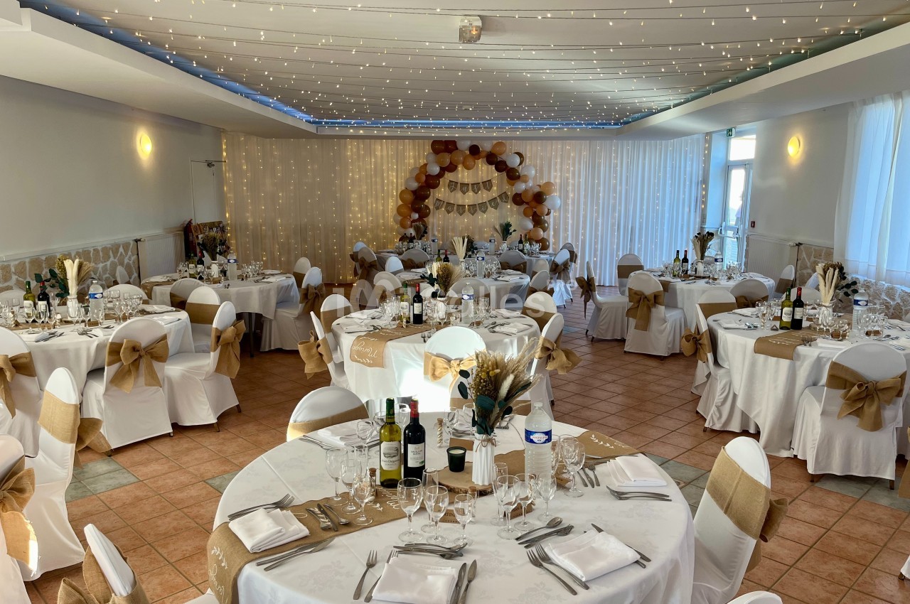 Salle de réception décorée avec des tables rondes, nappes blanches, chaises ornées de nœuds et guirlandes lumineuses au…