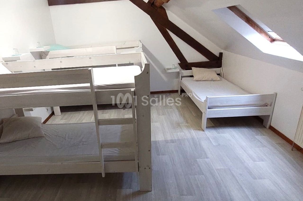 Chambre mansardée avec lits superposés en bois blanc, un lit simple, parquet clair et poutres apparentes.