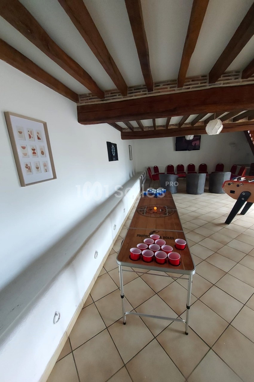 Table de beer-pong avec gobelets rouges dans une pièce à poutres apparentes, sol carrelé et baby-foot en arrière-plan.