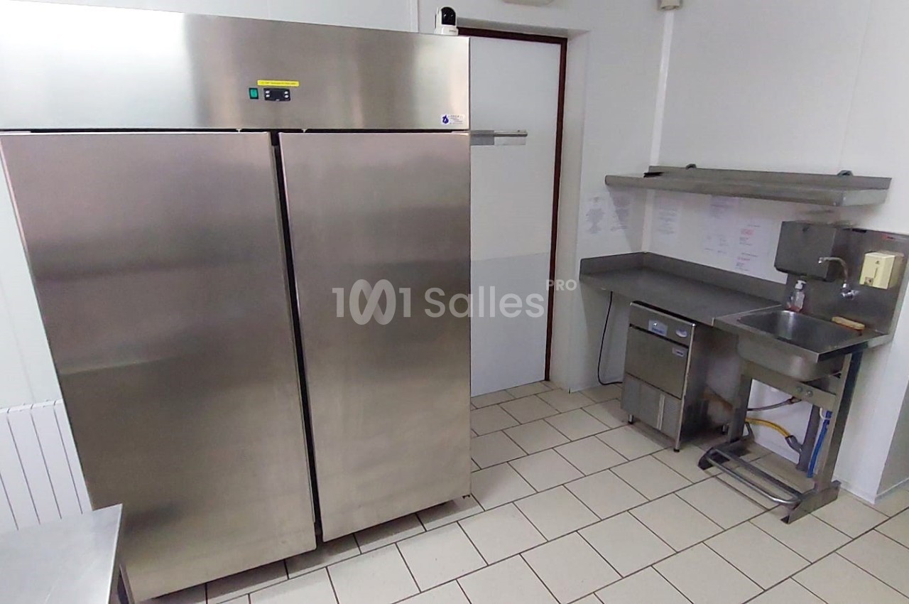 Cuisine professionnelle avec réfrigérateur en inox, évier, étagères murales et sol carrelé blanc.