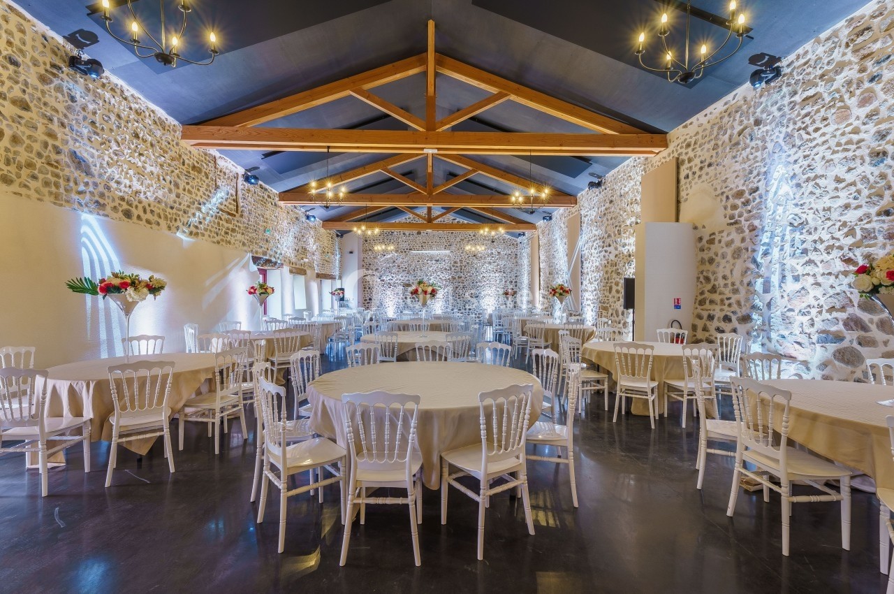 Salle de réception avec murs en pierre, poutres apparentes, tables rondes dressées et chaises blanches.
