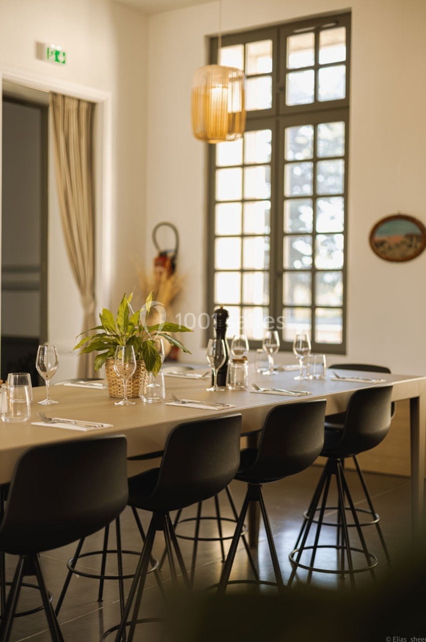 Table dressée avec des verres et couverts dans une salle lumineuse, décorée de plantes et d'une suspension en rotin.