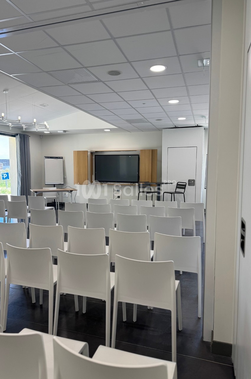 Salle de conférence lumineuse avec rangées de chaises blanches, tableau interactif et paperboard à l'avant.
