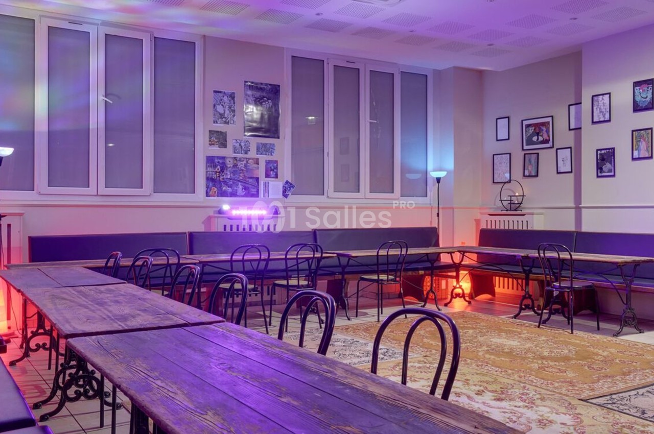 Salle lumineuse avec tables en bois, chaises noires, murs décorés de cadres et éclairage coloré.