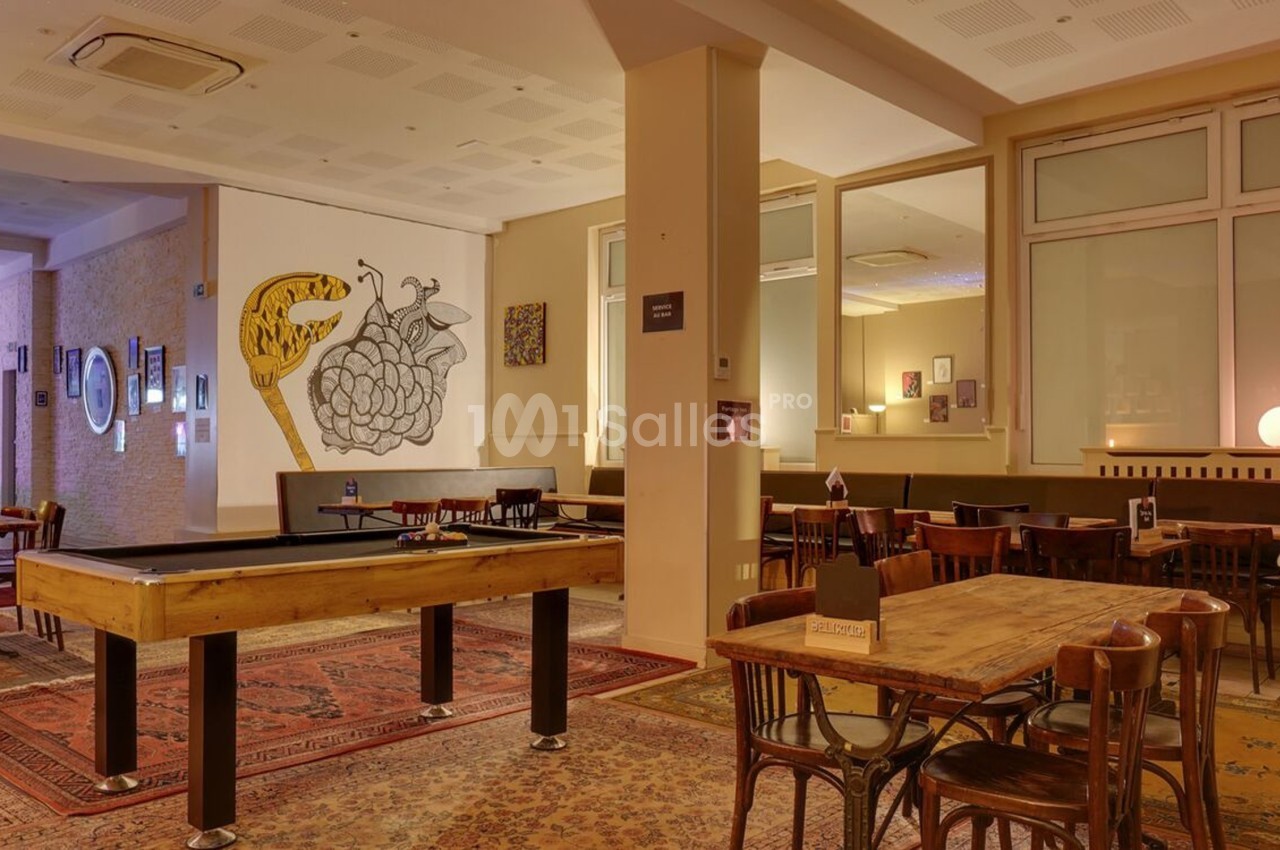Salle conviviale avec tables en bois, tapis colorés, baby-foot et décorations murales artistiques.
