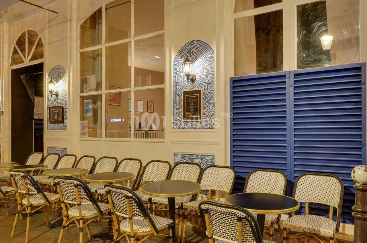 Façade d'un café avec terrasse extérieure, chaises en osier et tables rondes, éclairée par des appliques murales.