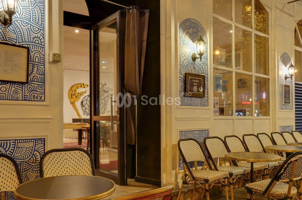 Façade d'un café avec terrasse extérieure, chaises en osier et tables rondes alignées, éclairage nocturne.