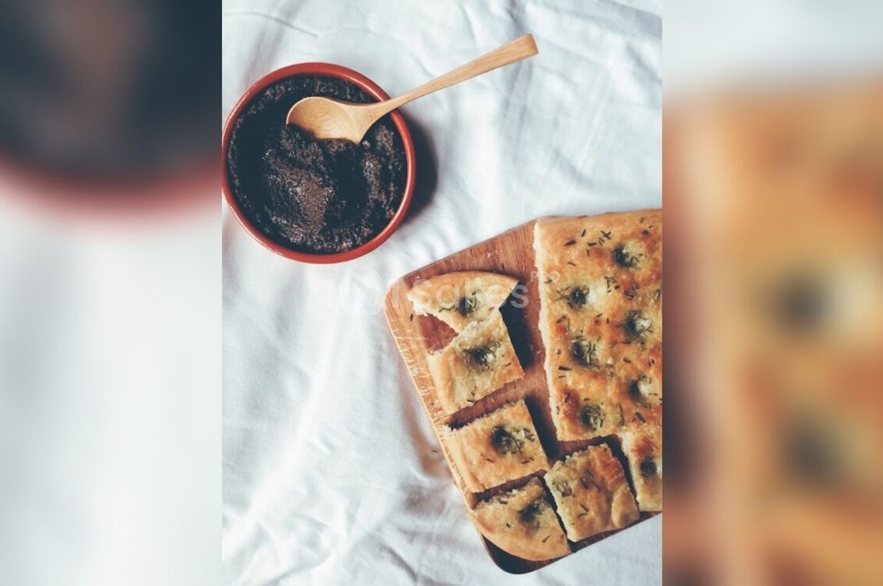 Focaccia coupée en morceaux sur une planche en bois, accompagnée d'un bol de tapenade et d'une cuillère en bois.