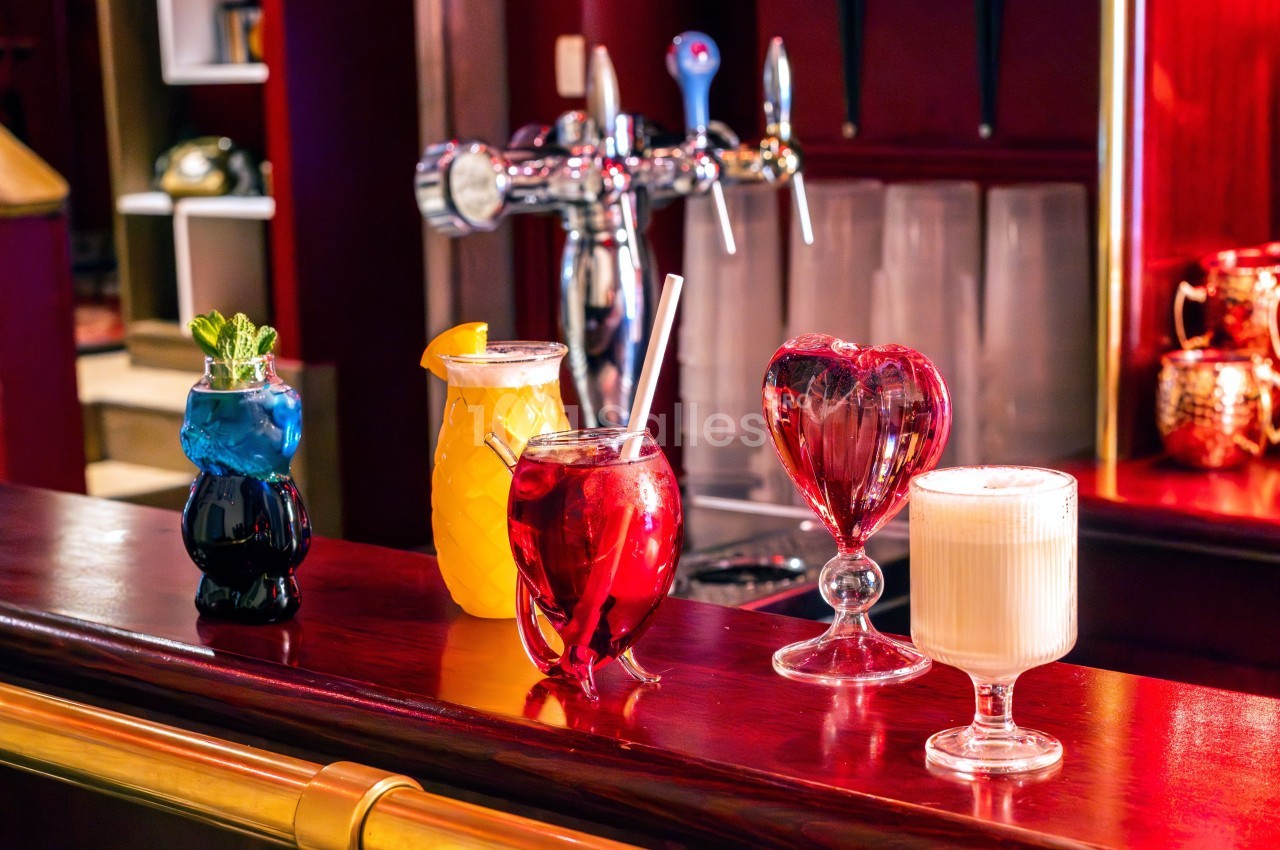 Quatre cocktails colorés servis dans des verres originaux sur un comptoir de bar en bois, avec des tireuses en arrière-plan.