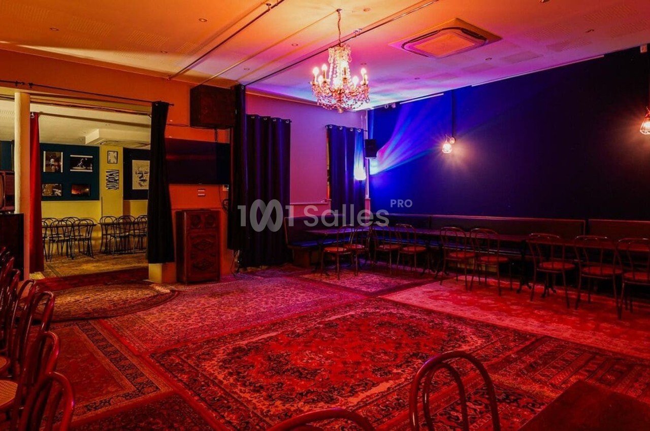 Salle avec tapis colorés, chaises alignées, murs sombres, éclairage tamisé et lustre central.