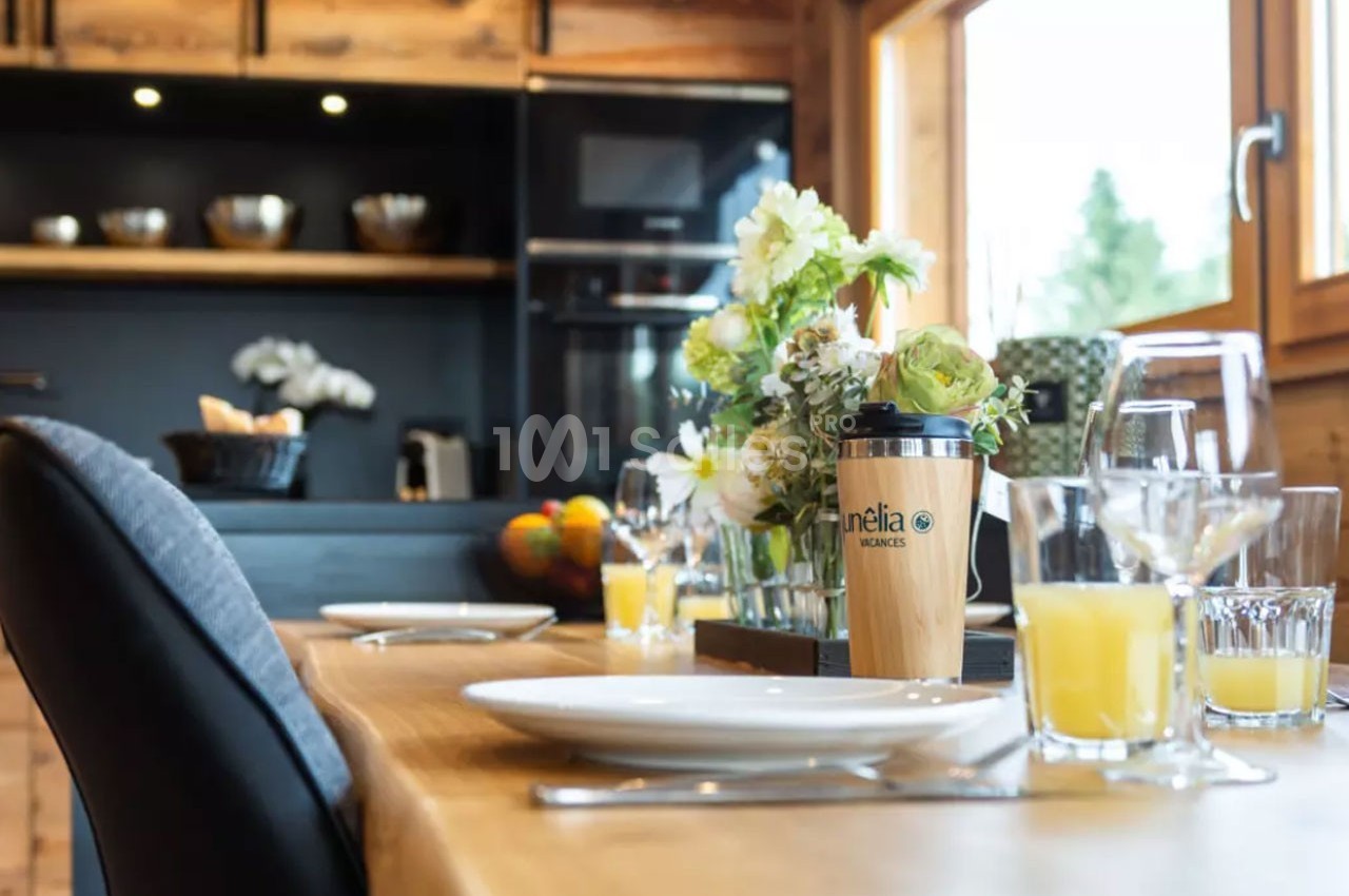 Table dressée avec des assiettes, verres de jus d'orange et un gobelet, dans une cuisine lumineuse en bois.