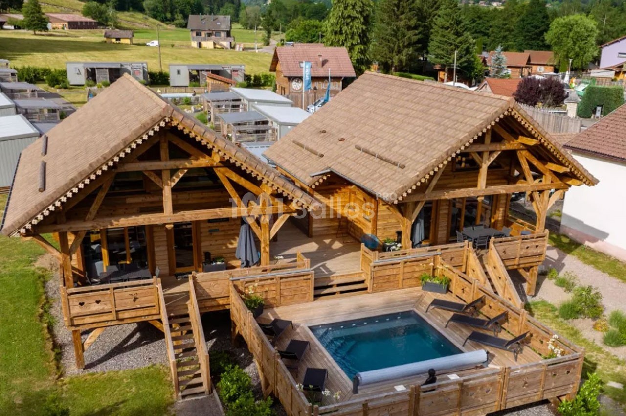 Deux chalets en bois avec une terrasse commune et un jacuzzi, situés dans un environnement verdoyant.