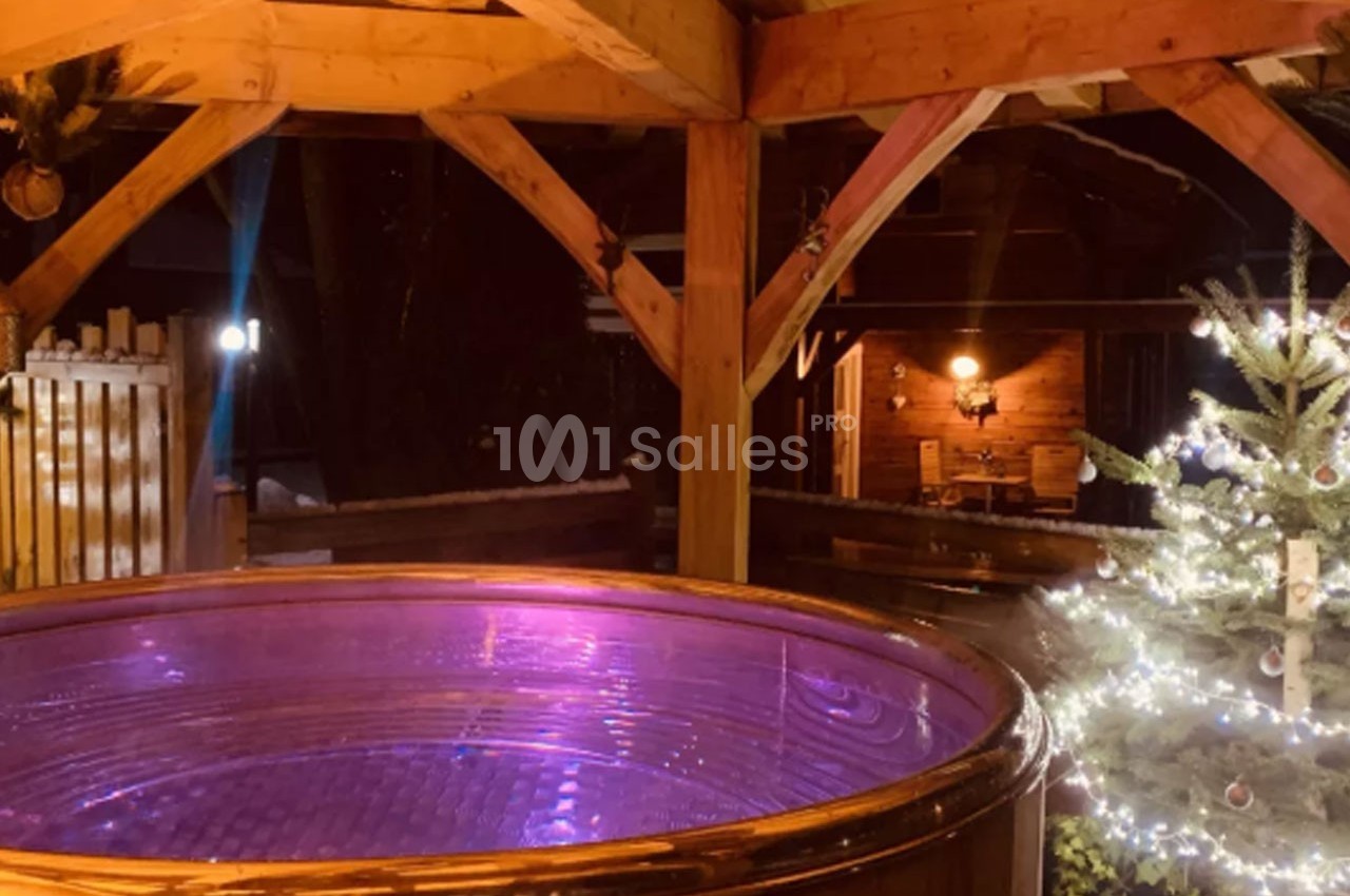 Bain à remous éclairé en violet sous une pergola en bois, avec un sapin décoré et un chalet en arrière-plan.