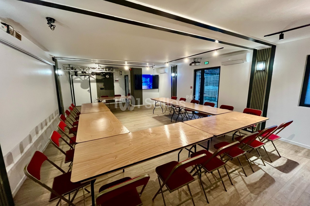 Salle de réunion lumineuse avec tables en bois disposées en U et chaises rouges, écran au mur en arrière-plan.