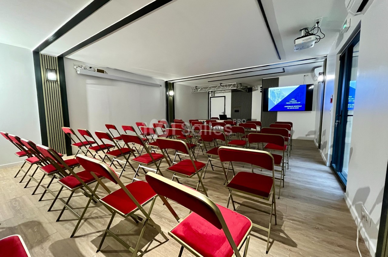 Salle de conférence avec des chaises rouges alignées face à un écran de projection et un tableau blanc.