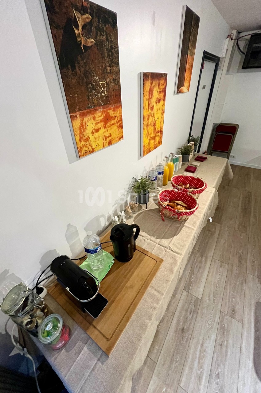 Table avec cafetière, carafe, paniers de viennoiseries, jus de fruits et décorations murales dans une pièce lumineuse.