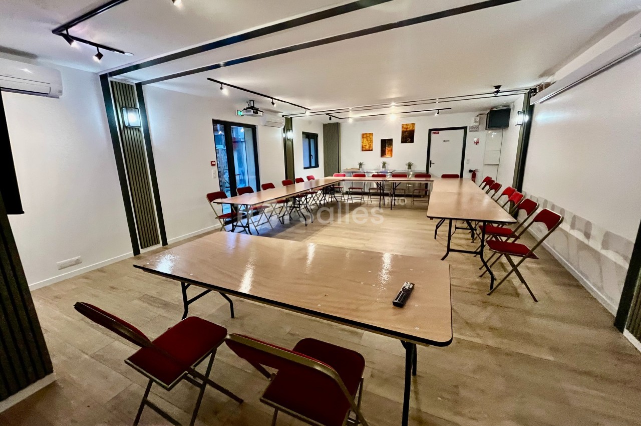 Salle de réunion lumineuse avec tables disposées en U, chaises rouges et équipement audiovisuel.