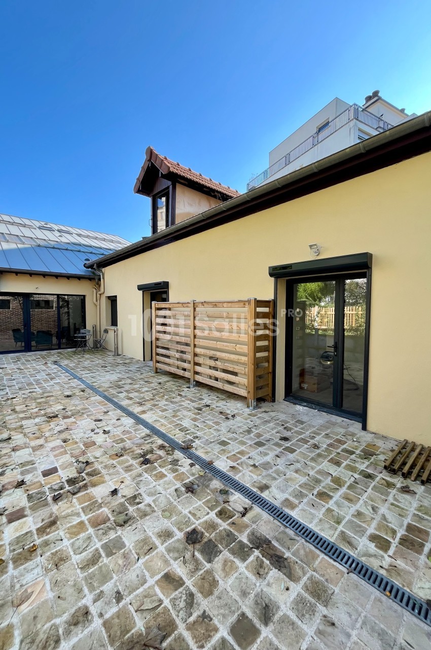 Cour pavée avec façade beige, grandes baies vitrées, clôtures en bois et ciel dégagé en arrière-plan.