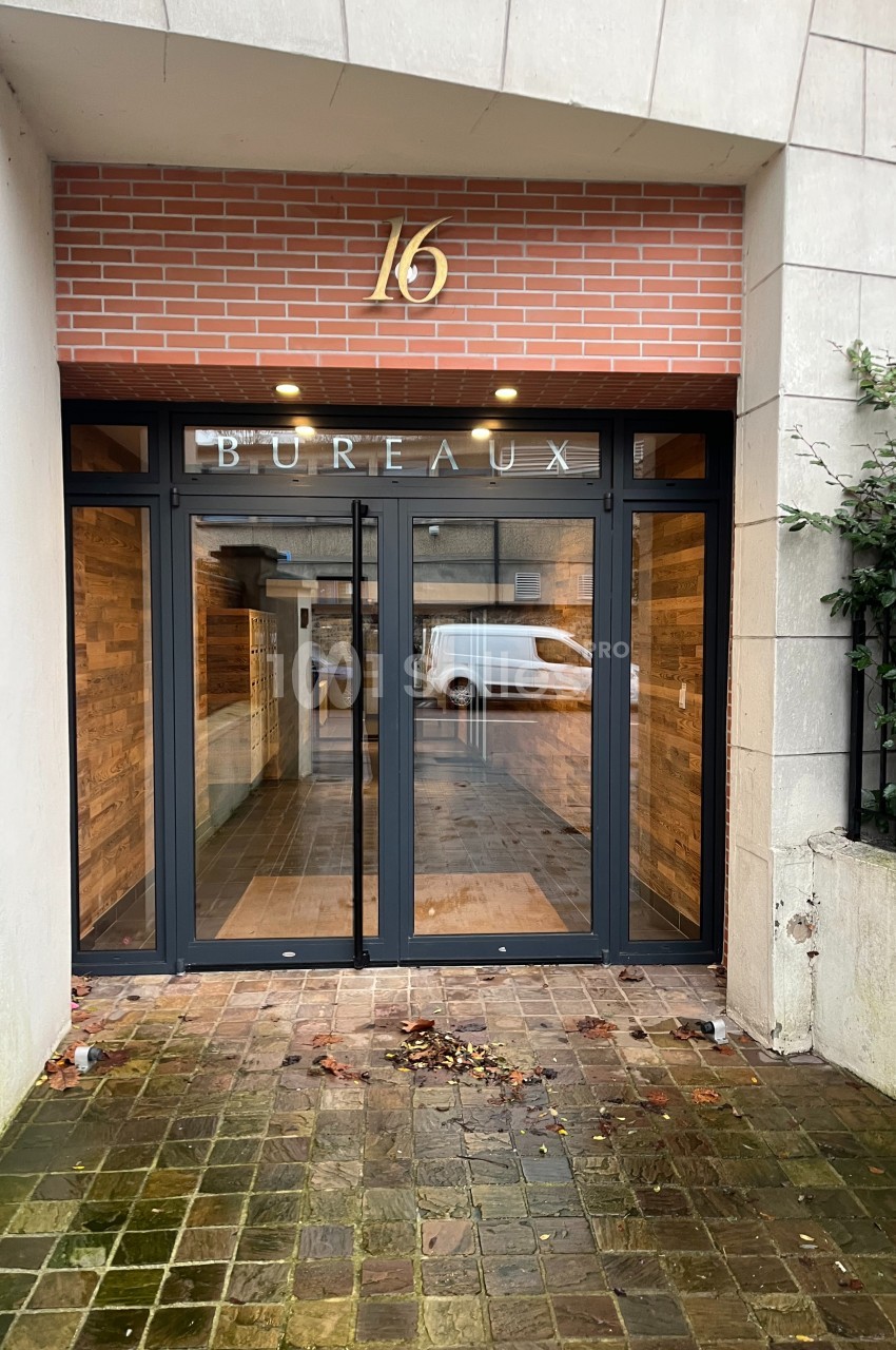 Entrée d'un immeuble avec porte vitrée, numéro 16 et inscription ’Bureaux’, sol pavé avec quelques feuilles.