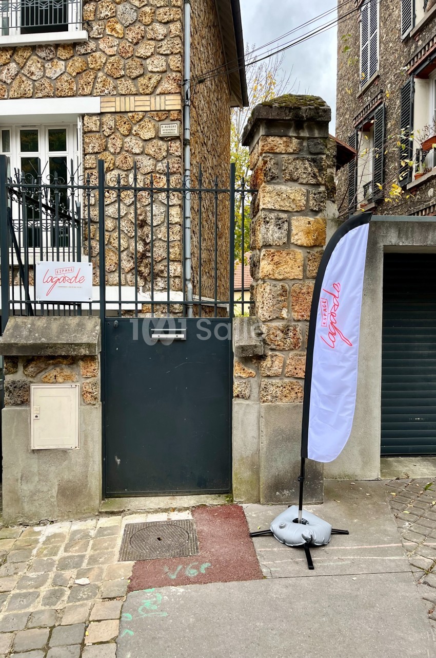 Portail noir en fer forgé encadré par un mur en pierre, avec une enseigne et un drapeau publicitaire à droite.