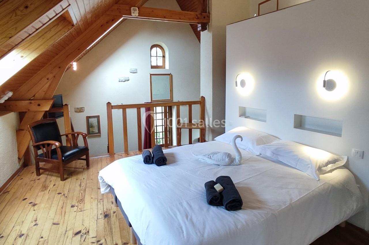 Chambre mansardée avec lit double, serviettes pliées, cygne en serviette, fauteuil et lumière naturelle.