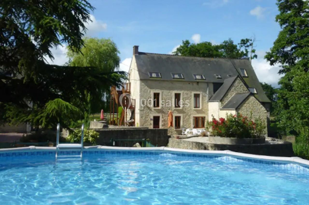 Maison en pierre avec toit en ardoise, entourée de verdure, vue depuis une piscine extérieure sous un ciel dégagé.
