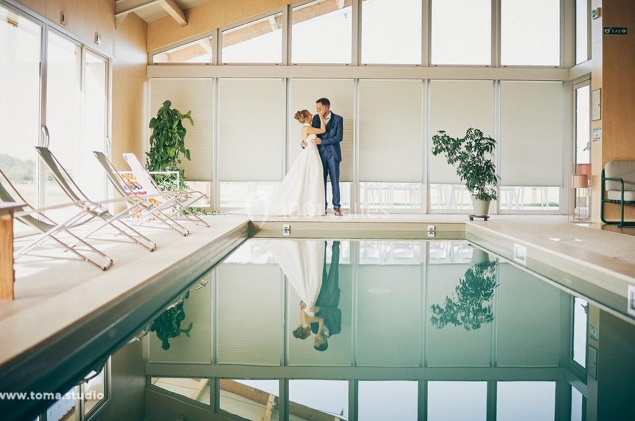 Un couple en tenue de mariage se tient près d'une piscine intérieure entourée de chaises longues et de plantes.