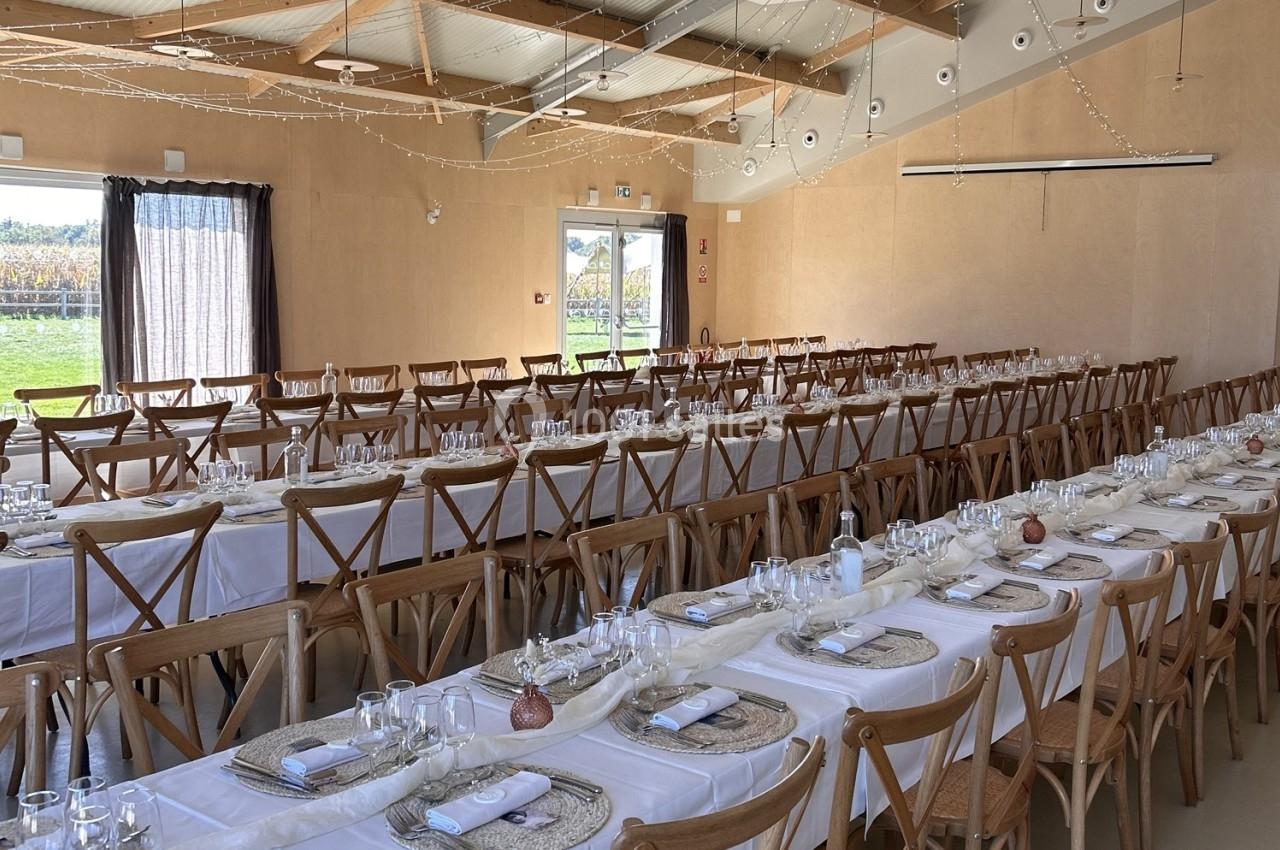 Salle de réception avec de longues tables dressées, nappes blanches, chaises en bois et décorations simples.