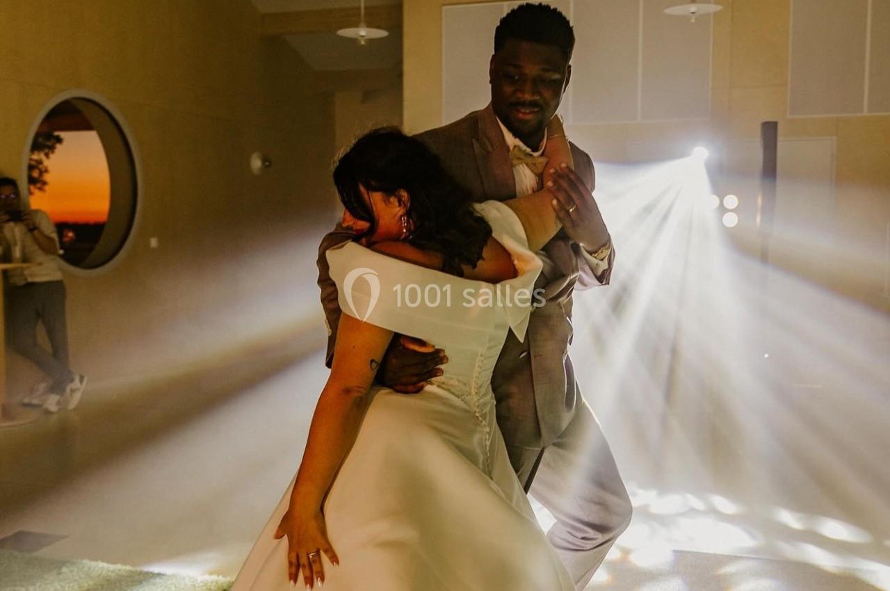 Un couple danse dans une salle éclairée par des projecteurs, avec un coucher de soleil visible par une fenêtre ronde.