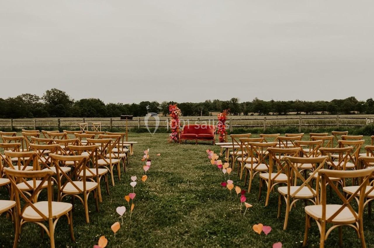 Chaises en bois disposées en allées face à un canapé rouge décoré de fleurs, dans un champ en plein air.